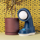 The Nessi table lamp in Navy sits next to a plant, in front of yellow floral wallpaper