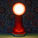 A head-on view of the Nessi table lamp in a dark room with floral wallpaper behind it.