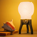 The glowing Visitor table lamp in Olive, next to an art book with a seashell on top.